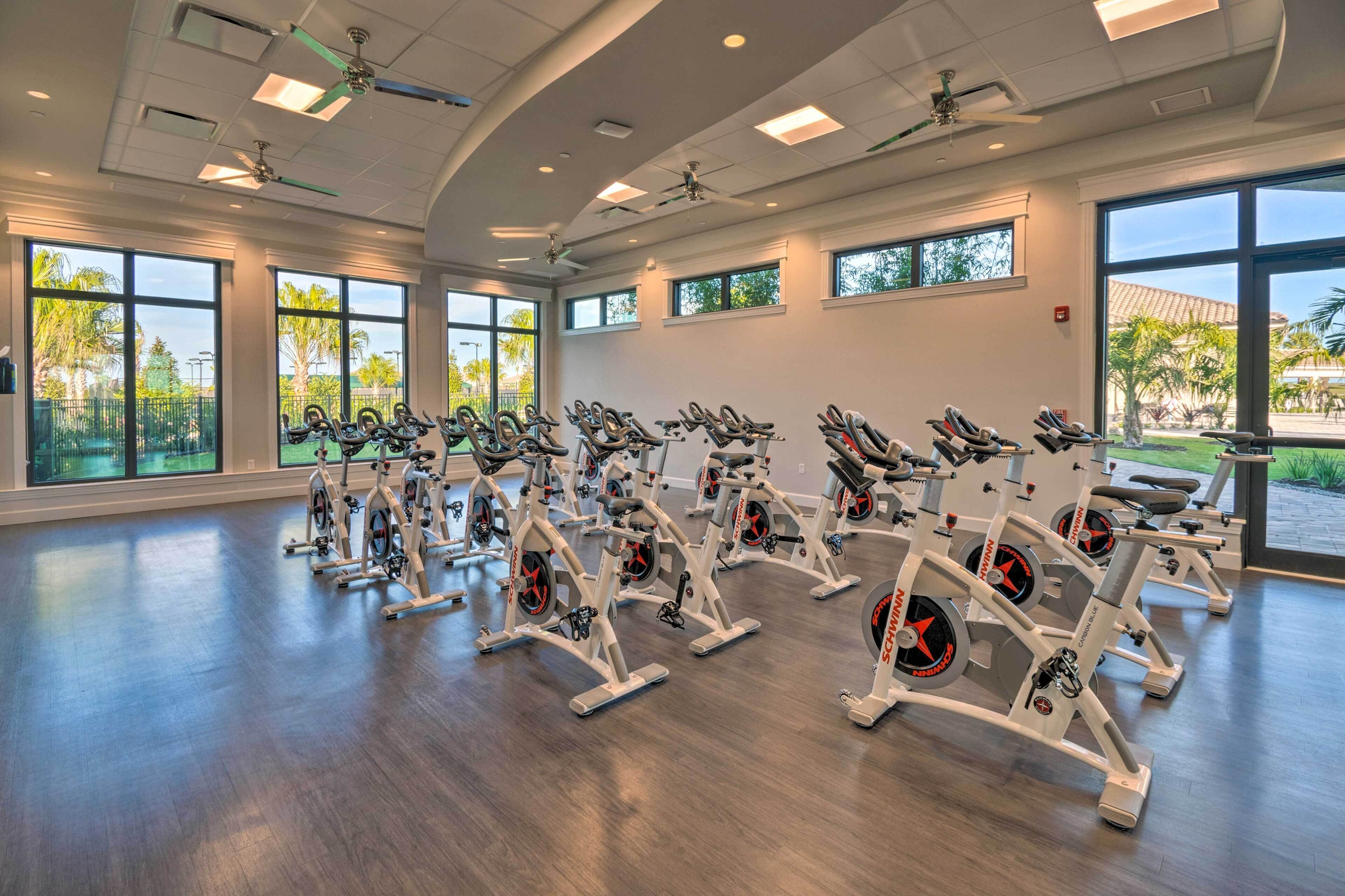 A gym with stationary bikes arranged in rows, large windows, and ceiling fans.