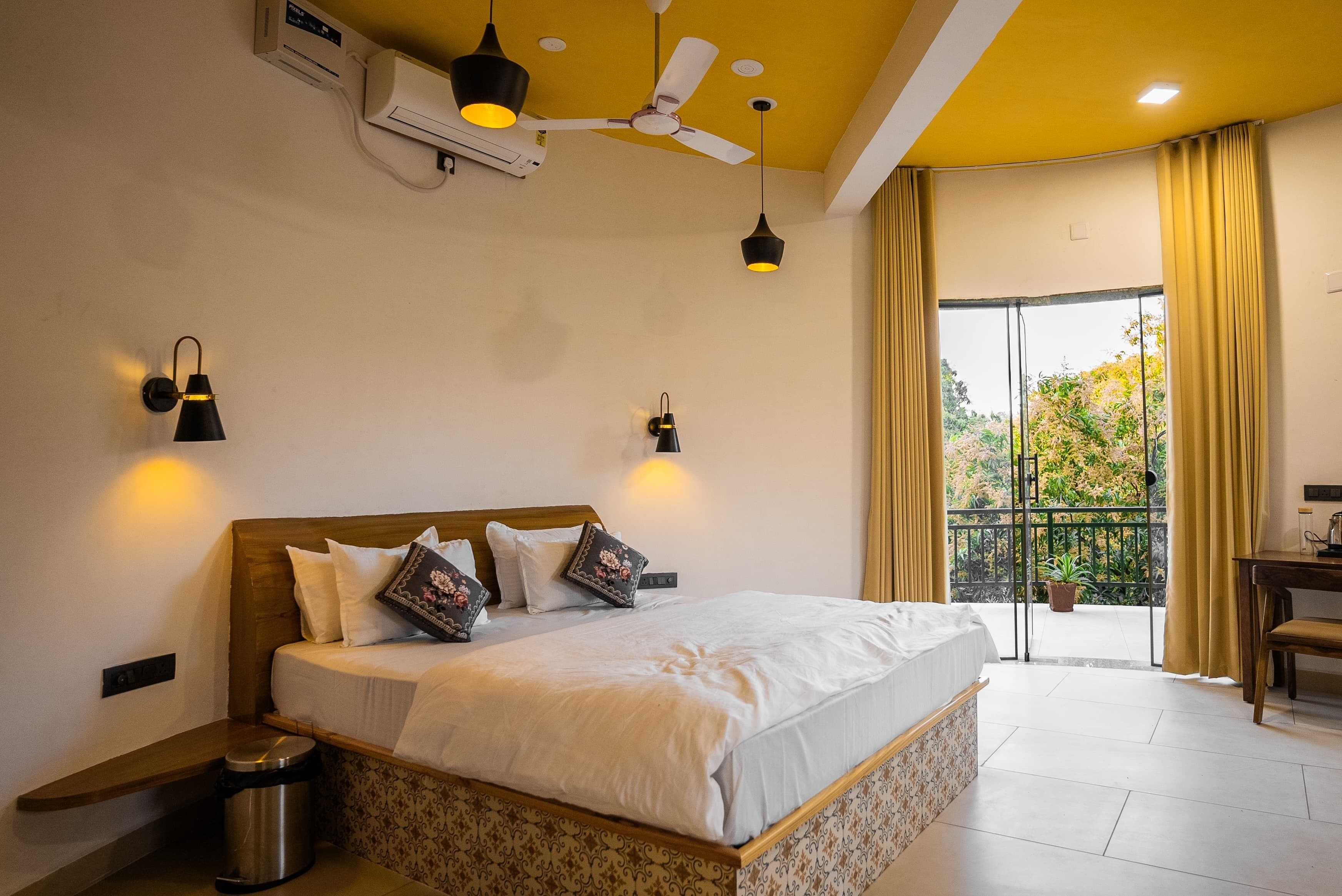 A hotel room with a bed, bedside table, trash bin, ceiling fan, wall-mounted lights, and a view of the outdoors through a large window.