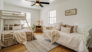 Un dormitorio con una cama litera, una cama individual, un ventilador de techo, una mesita de noche con lámpara y una pared decorada con una cita enmarcada.