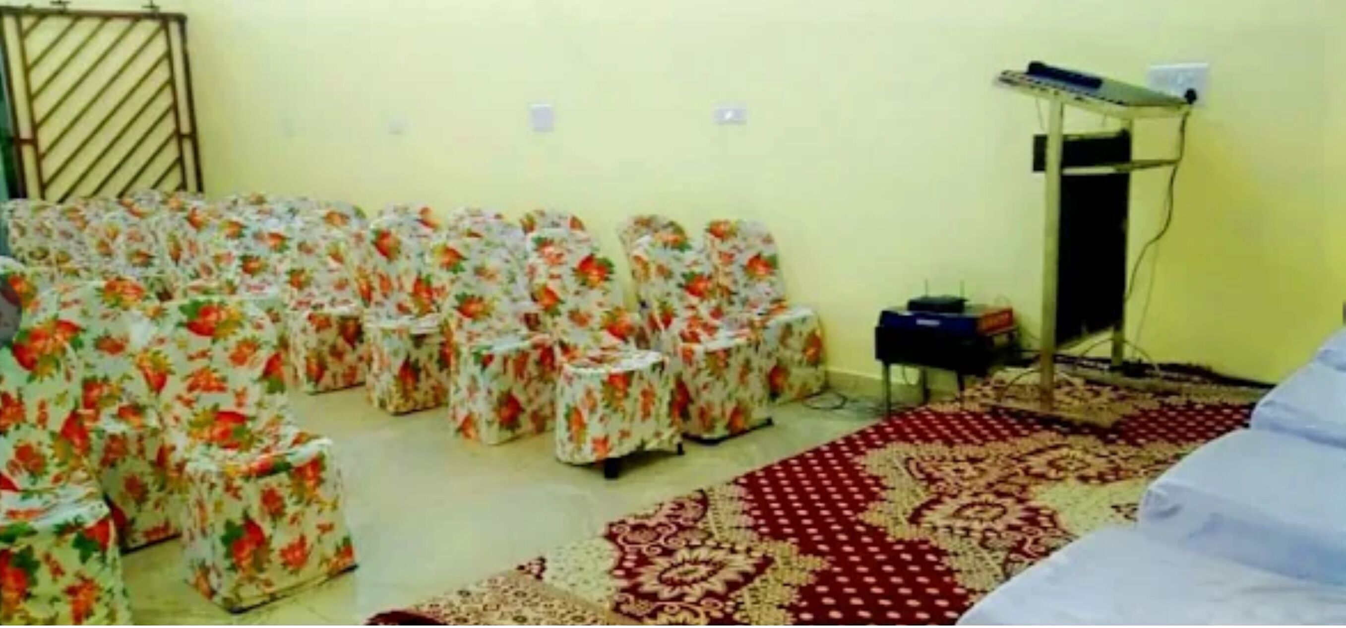 A room with multiple floral-patterned chairs arranged in rows, a small table, a bed, a patterned carpet, and a medical equipment stand.