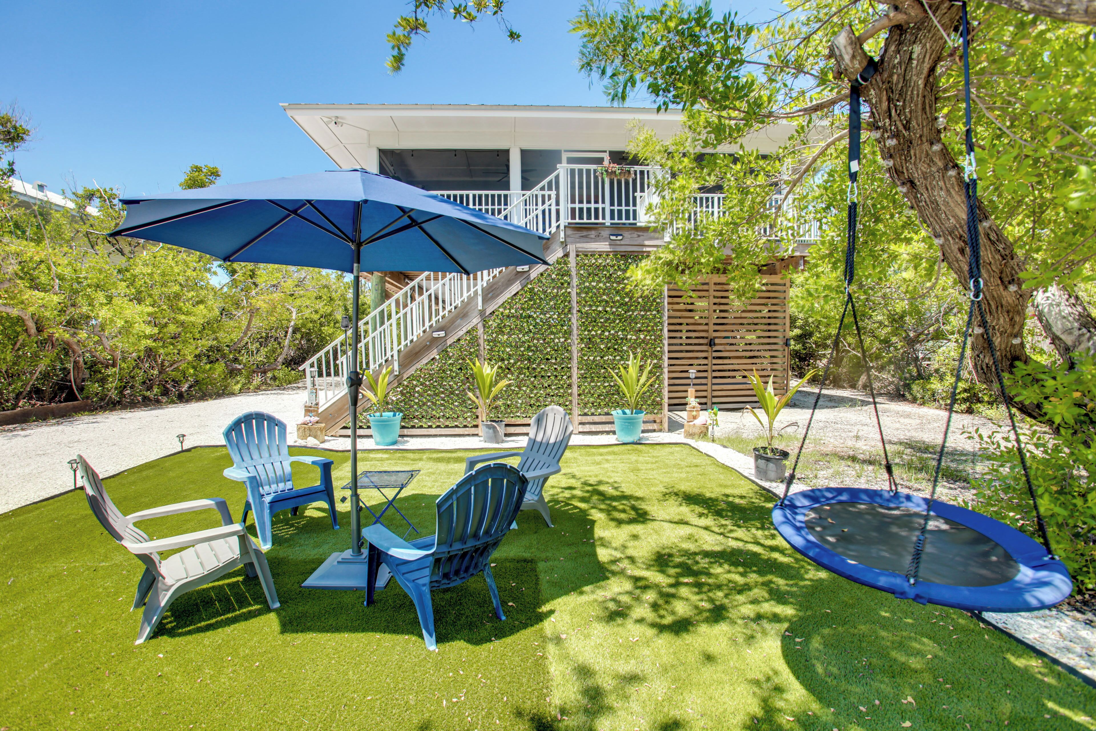 Un patio trasero con un trampolín, dos sillas Adirondack azules, una mesa y una sombrilla.