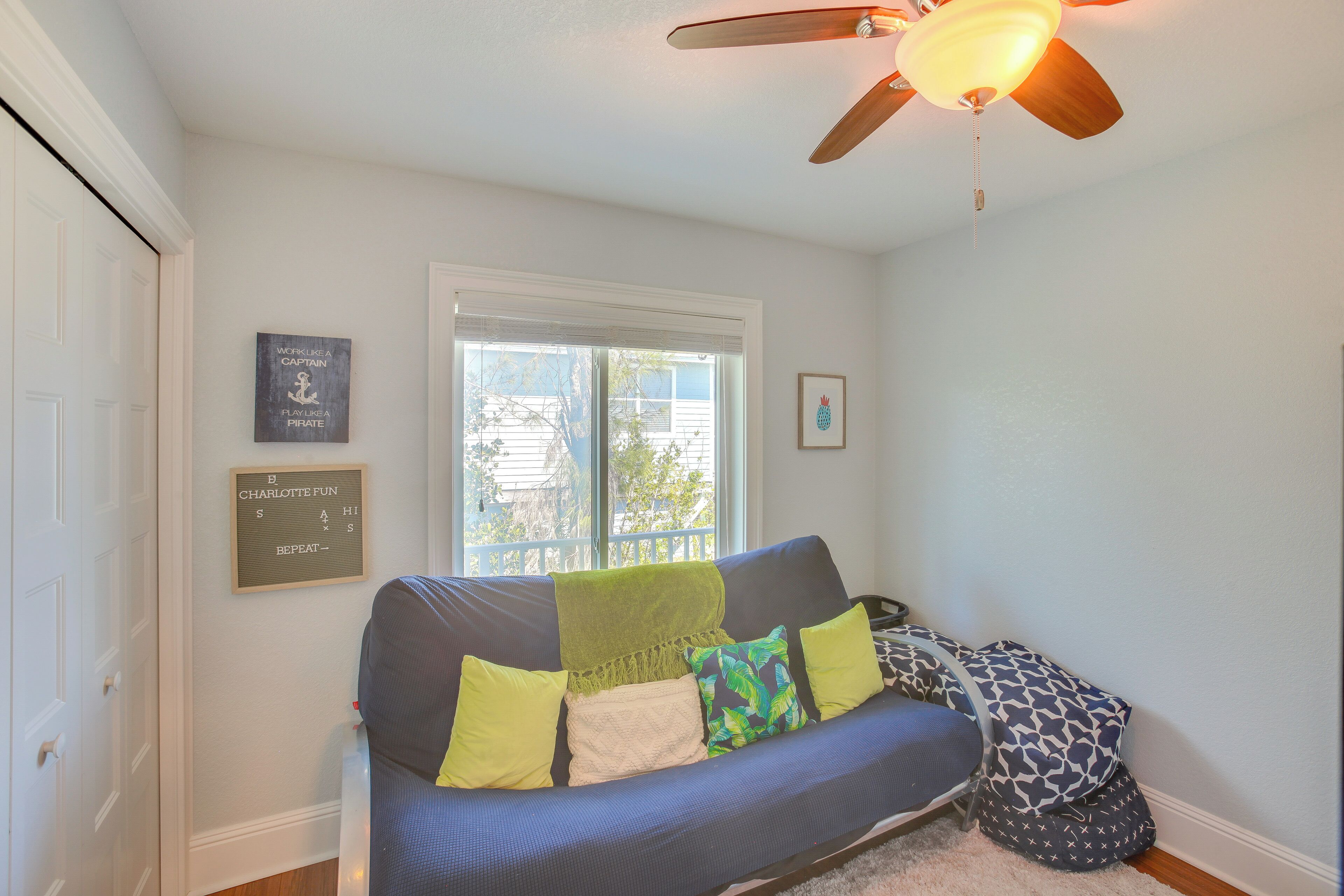A living room with a blue sofa, green and yellow cushions, a ceiling fan, and a window with blinds.