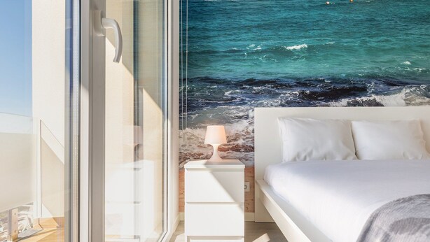A bedroom with a large window overlooking a beach, a bedside table with a lamp, and a bed with white linens.