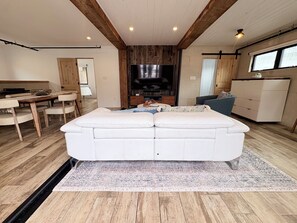 A modern living room with a large white sofa, a flat-screen TV, wooden beams on the ceiling, and a dining area with a table and chairs. - Cali beach living! Modern 2 BR home. Pet friendly. Walk to town, beaches, harbor (Dana Point)