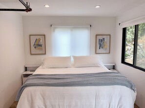Une chambre à coucher moderne, avec un lit, deux tables de chevet, une fenêtre donnant sur un beau paysage et deux œuvres d’art encadrées accrochées aux murs.