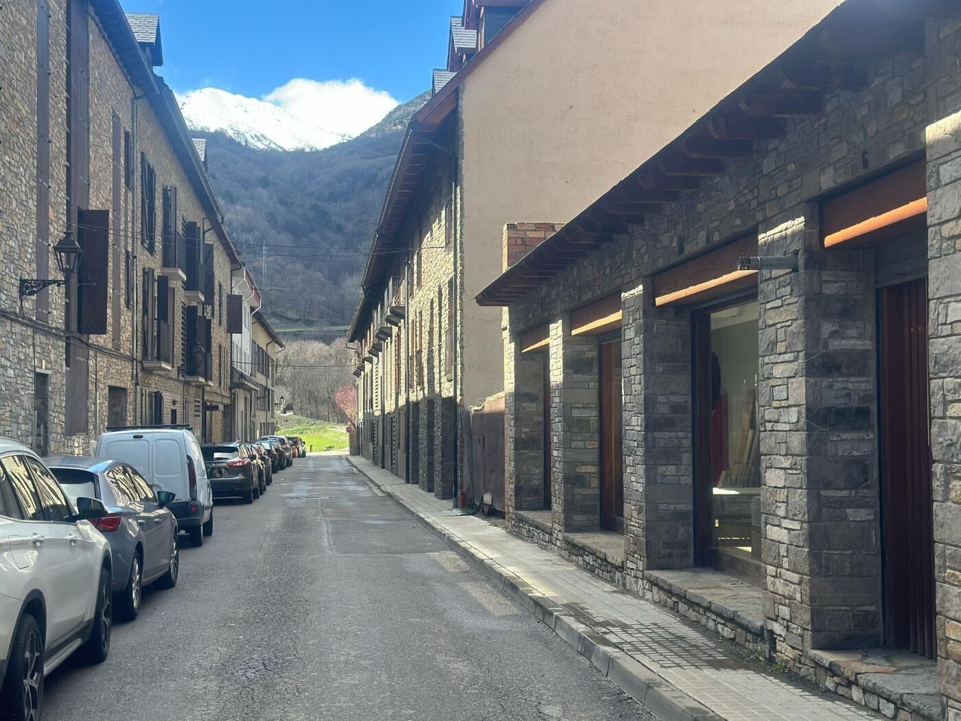 A street lined with stone buildings, parked cars, and a mountain in the background.