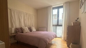 Un dormitorio con cama, mesita de noche, radiador, ventana con cortinas y suelo de madera.