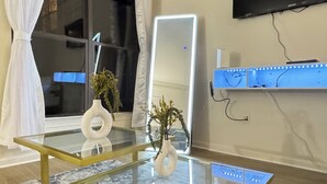 Una sala de estar moderna con una mesa de centro de vidrio, un espejo grande con luces LED, un televisor de pantalla plana montado en la pared y un jarrón decorativo con plantas.