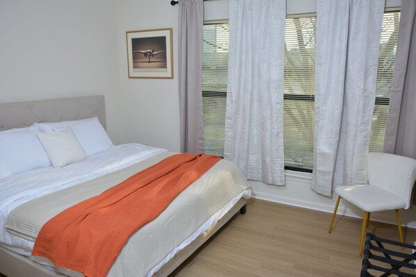A neatly made bed with a white headboard, a framed picture on the wall, and a window with white curtains.