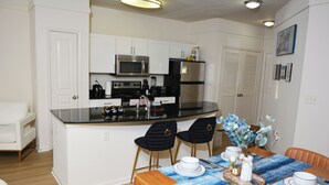 Cocina moderna con armarios blancos, electrodomésticos de acero inoxidable y una zona de comedor con mantel azul y platos blancos.