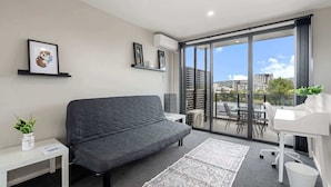 En moderne lejlighed med en grå sofa, et lille hvidt bord, et hvidt skrivebord og en balkon med udsigt til bygninger og grønne områder.