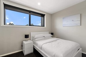 A bedroom with a bed, a nightstand, a window with a view of the sky, and a painting on the wall.