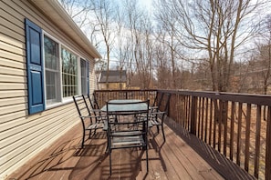 Une terrasse en bois avec une table et des chaises, une rambarde et une vue sur des arbres et une maison au loin.