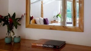 Un miroir à cadre en bois reflète une pièce où l’on aperçoit un lit, des oreillers et une fenêtre. Un livre intitulé « NGORONGORO » est posé sur une table en bois, à côté du miroir.