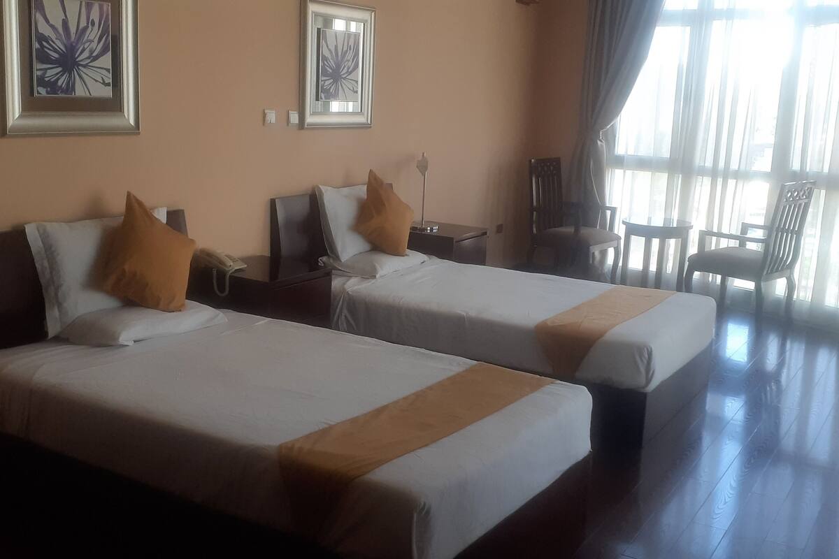 A hotel room with two beds, a desk, a chair, and large windows.