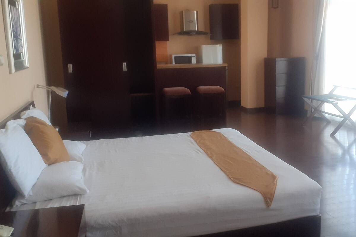 A hotel room with a bed, a desk, a chair, a TV, and a kitchenette.