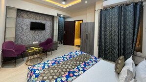 A modern bedroom with a bed, a TV mounted on the wall, a ceiling fan, and a seating area with chairs. - Kashi Kutir (Varanasi)
