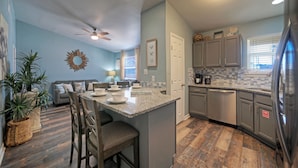 A kitchen with a central island, bar stools, a ceiling fan, and a refrigerator. - Stylish 2BR Coastal Getaway w/ Pool & Prime Location | Lazy Fun (Port Aransas)