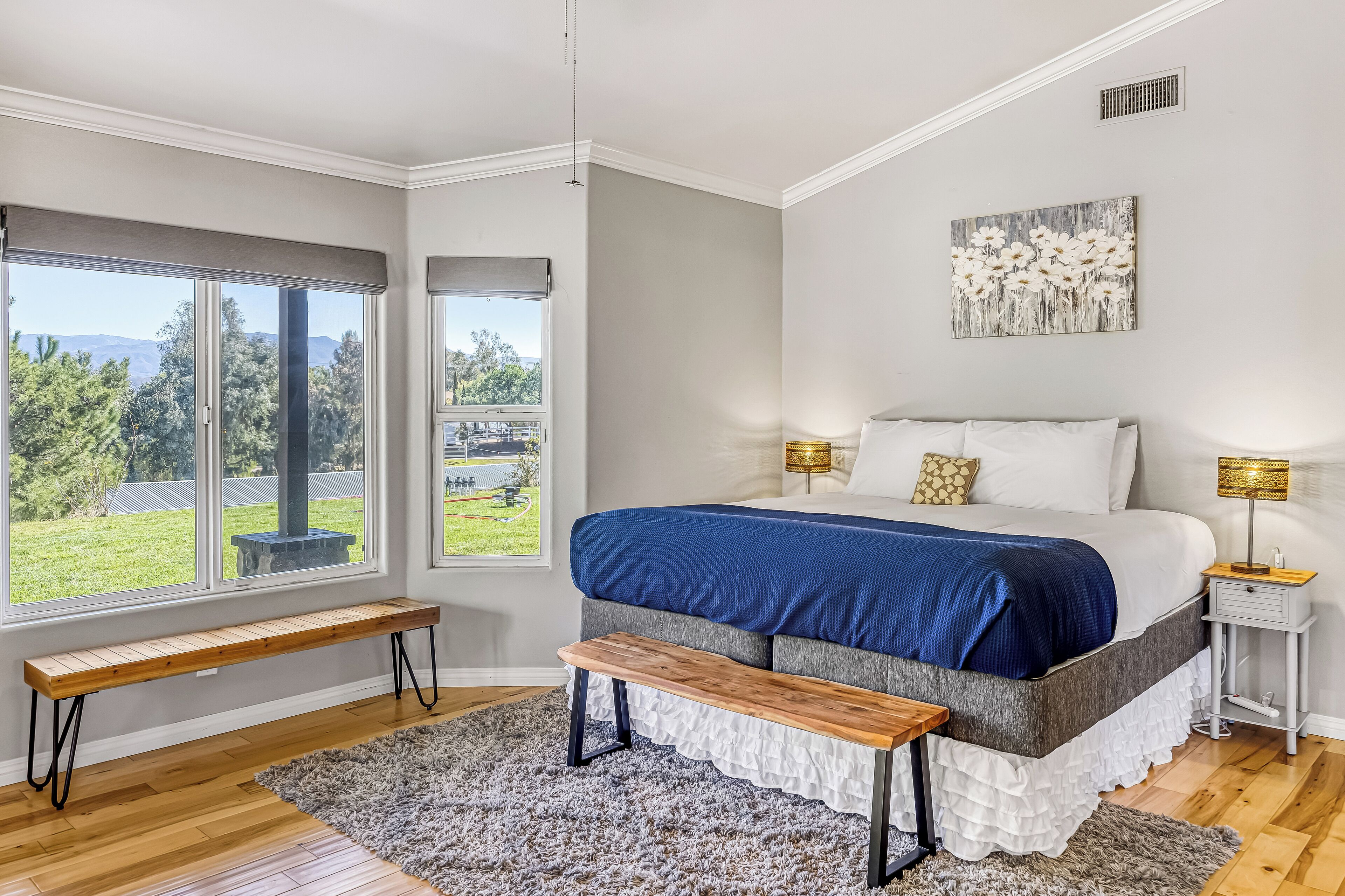 A bedroom with a large bed, a bench, a view of the outdoors, and a painting on the wall.