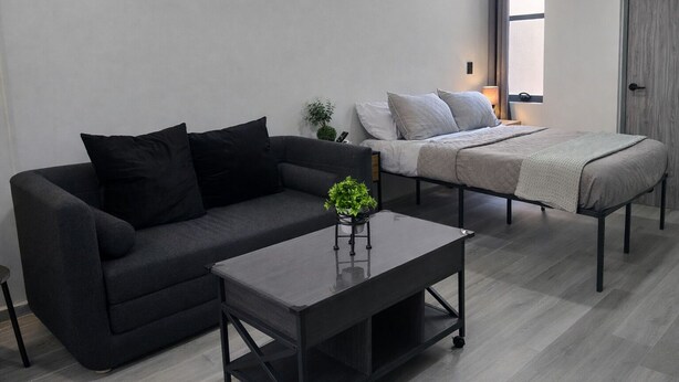A modern living room with a dark grey sofa, a matching coffee table, and a bed with a grey duvet.