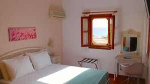 A bedroom with a bed, a desk, a chair, a window with a view, and a wall-mounted air conditioner. - Cavo Grosso Bungallows (Mavrovouni)