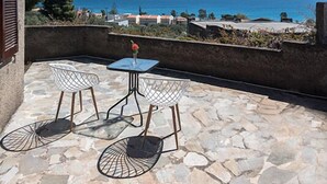 A patio with stone flooring, two white wire chairs, and a small table. A view of the sea and buildings is visible in the background. - Cavo Grosso Bungallows (Mavrovouni)
