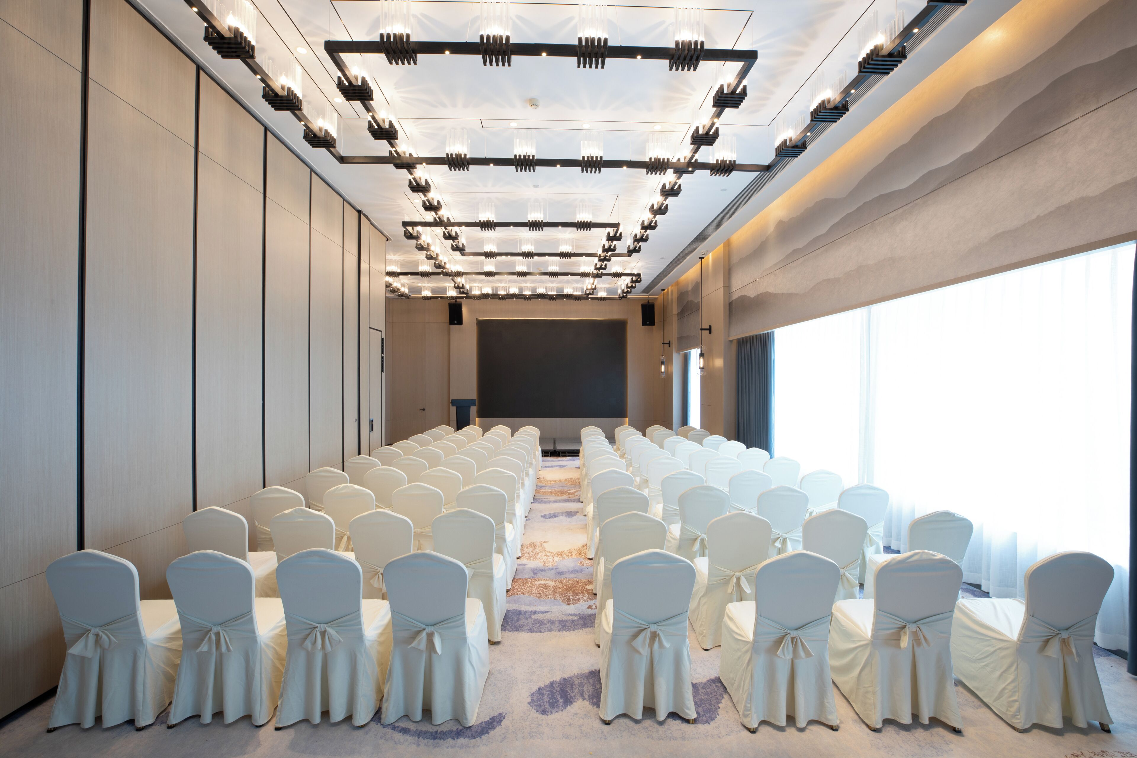 A conference room with rows of white chairs, a large screen at the front, and a modern ceiling design.