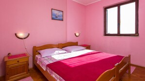 Dormitorio con decoración rosa, una cama de madera, dos mesitas de noche, una mesilla auxiliar, una ventana y un cuadro en la pared.