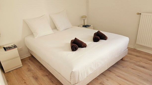 A neatly made bed with white linens, two rolled-up towels, a bedside table with a lamp, and a book.