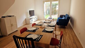 A dining area with a wooden table set for four, surrounded by red chairs. A television is mounted on the wall, and a blue sofa is visible in the background.