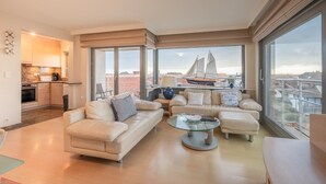 Un salon moderne avec un ensemble de canapés, une table basse en verre et une vue sur un voilier, visible à travers de grandes fenêtres.