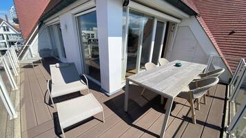 Een overdekt terras buiten met een tafel en stoelen, een schuifpui en uitzicht op omliggende gebouwen.