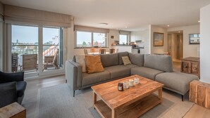 Een ruime woonkamer met een grote bank, een houten bijzettafeltje en stadszicht door grote ramen.