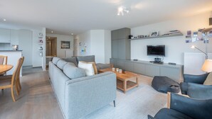 Een moderne woonkamer met een grijze bank, een houten salontafel en een televisie aan de muur.