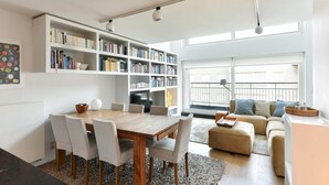 Ein modernes Wohnzimmer mit einem Holzesstisch, weißen Stühlen, einem beige gefärbten Sofa und einem Bücherregal voller Bücher.