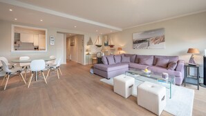 A modern living room with a purple sectional sofa, a glass coffee table, and a dining area with white chairs and a glass table. - Apartment in Duinbergen near Sailing School (KNOKKE-HEIST)