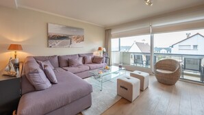A modern living room with a large sofa, a glass coffee table, and a wicker chair. There is a painting on the wall and a view of buildings through large windows. - Apartment in Duinbergen near Sailing School (KNOKKE-HEIST)