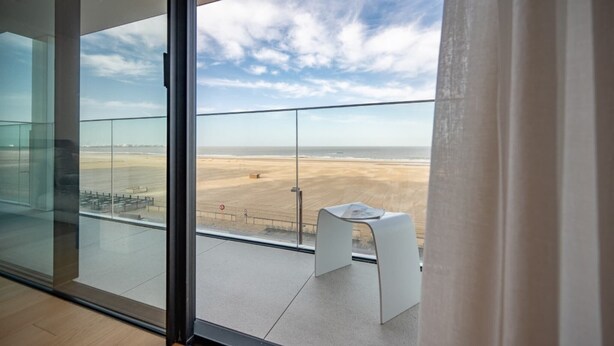 A modern room with a large glass door leading to a balcony with a view of a beach and distant cityscape. - Apartment in Knokke with Sea View (KNOKKE-HEIST)