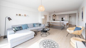 Una sala moderna con sofá, mesa de centro y silla de mimbre. Se observa una cocina al fondo.