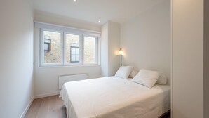 Un dormitorio con cama, dos ventanas, un radiador y una lámpara fijada a la pared.