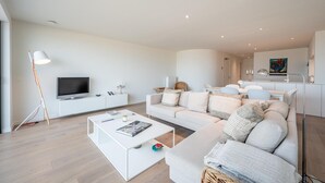 Una sala de estar moderna con un sofá blanco, una mesa de centro, un televisor de pantalla plana y un comedor con una mesa y sillas blancas.