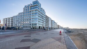 A modern multi-story hotel with a prominent corner design, a paved walkway, and a sandy beach area. - Apartment in Knokke near Casino and Beach (KNOKKE-HEIST)