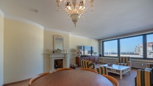 A spacious living room with a large wooden dining table, striped armchairs, a fireplace, and a large window offering a view of the city. - Apartment in Knokke near Casino and Beach (KNOKKE-HEIST)