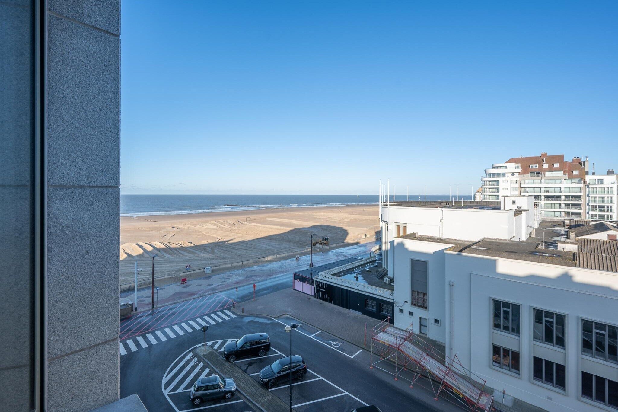 Blick aus einem Fenster auf einen Parkplatz mit Autos, ein Gebäude mit flachem Dach und einen Strand in der Ferne.