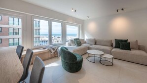 A modern living room with a sofa, chairs, a coffee table, and large windows offering a city view.