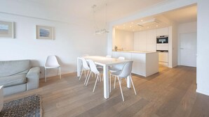 Une cuisine moderne avec des meubles blancs, un coin repas comprenant une table et des chaises blanches, et un canapé gris.