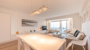 Un salon moderne doté d’une grande fenêtre, un coin repas avec une longue table et des chaises, et un téléviseur posé sur une table basse.