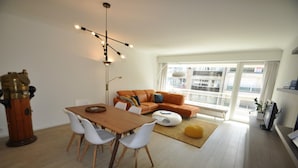 Un salon moderne avec une table à manger en bois, des chaises blanches, un canapé orange et un pouf jaune.