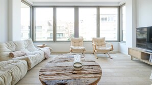 Ein geräumiges Wohnzimmer mit großem Fenster, einer Couch, zwei Sesseln, einem Couchtisch und einem Fernseher.