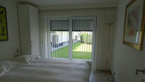 Un dormitorio con una cama grande, un ventanal corredero a un balcón, una lámpara de pie y un cuadro en la pared.